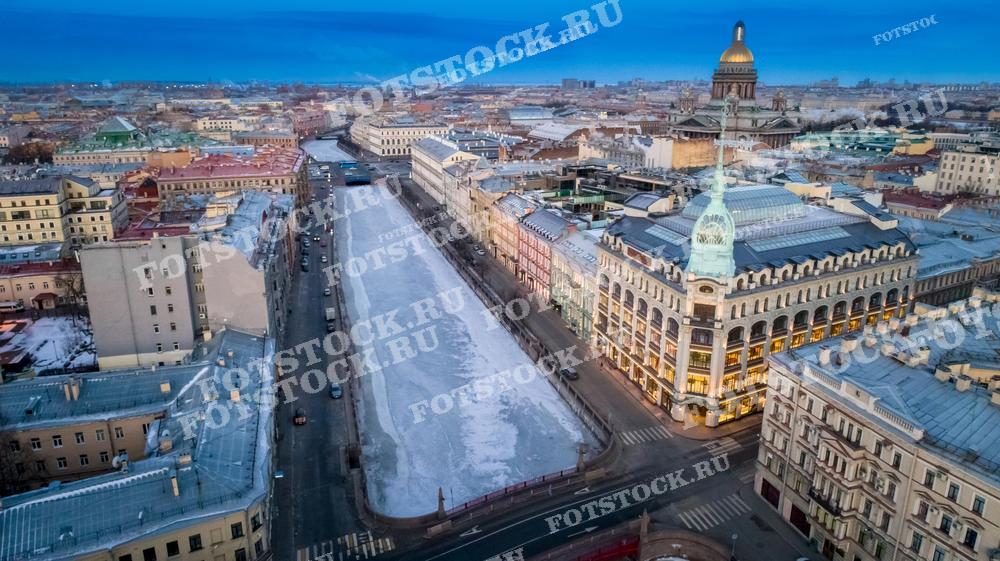На фото изображена панорама центра Санкт-Петербурга, снятая с высоты в зимний день. Вдалеке можно увидеть золотистый купол Исаакиевского собора, который гармонично вписывается в городской пейзаж. Фотография иллюстрирует городскую жизнь и архитектуру. Она может быть использована для туристических каталогов, культурно-исторических публикаций и как декоративный элемент для офисных и жилых пространств.